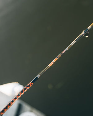 Close-up of a Camo Dock Runner Spinning Combo with orange and black patterned sections and a clear shaft, shown against a dark blurred background.