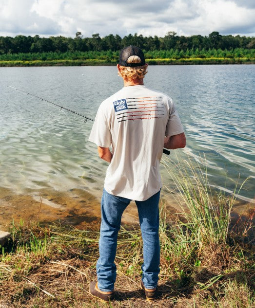 Man fishing by a lake with a cap and Ugly Stik branded  t-shirt on