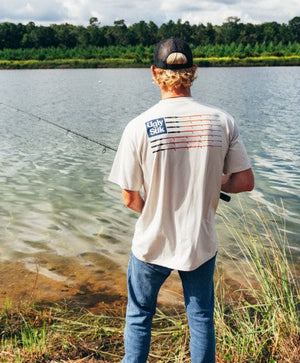 Man fishing by a lake with a cap and Ugly Stik branded  t-shirt on