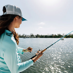 A woman in a light blue long-sleeve shirt, blue shorts, and a white cap is holding an Ugly Stik Carbon Inshore bent fishing rod over a body of water with a tree-lined shore in the background.