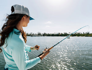 A woman in a light blue long-sleeve shirt, blue shorts, and a white cap is holding an Ugly Stik Carbon Inshore bent fishing rod over a body of water with a tree-lined shore in the background.