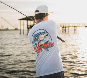 A man wearing a white t-shirt with a large blue and red fish graphic and Ugly Stik logo on the back, standing on a dock by the water at sunset.