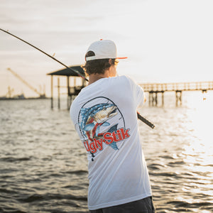 A man wearing a white t-shirt with a large blue and red fish graphic and Ugly Stik logo on the back, standing on a dock by the water at sunset.