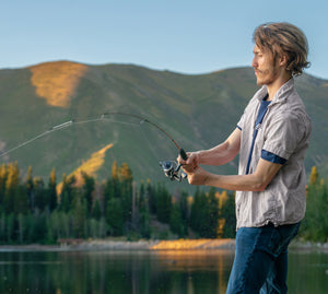 A man stands outdoors in front of mountains and trees, holding a Dock Runner Camo Spinning Combo. He is wearing a light gray short-sleeve button-up shirt over a blue undershirt.