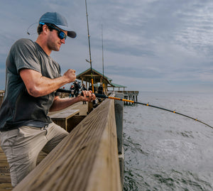 Man fishing off a pier with a Bigwater bent rod over water. He is wearing a green t-shirt, blue hat and sunglasses. 