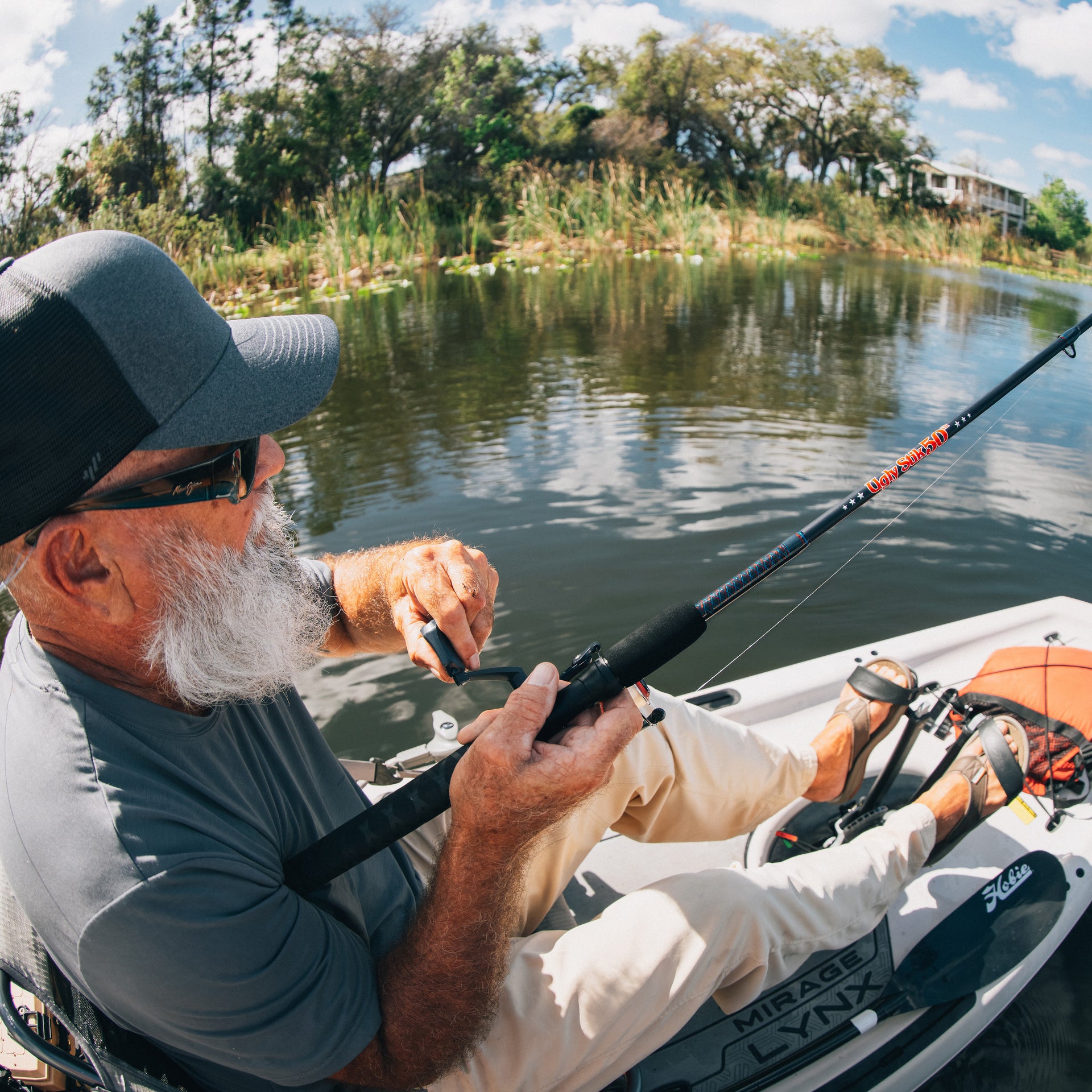 Man fishing from a kayak with an Ugly Stik 50th Anniversary Spinning Combo on a calm lake with trees and a house in the background.