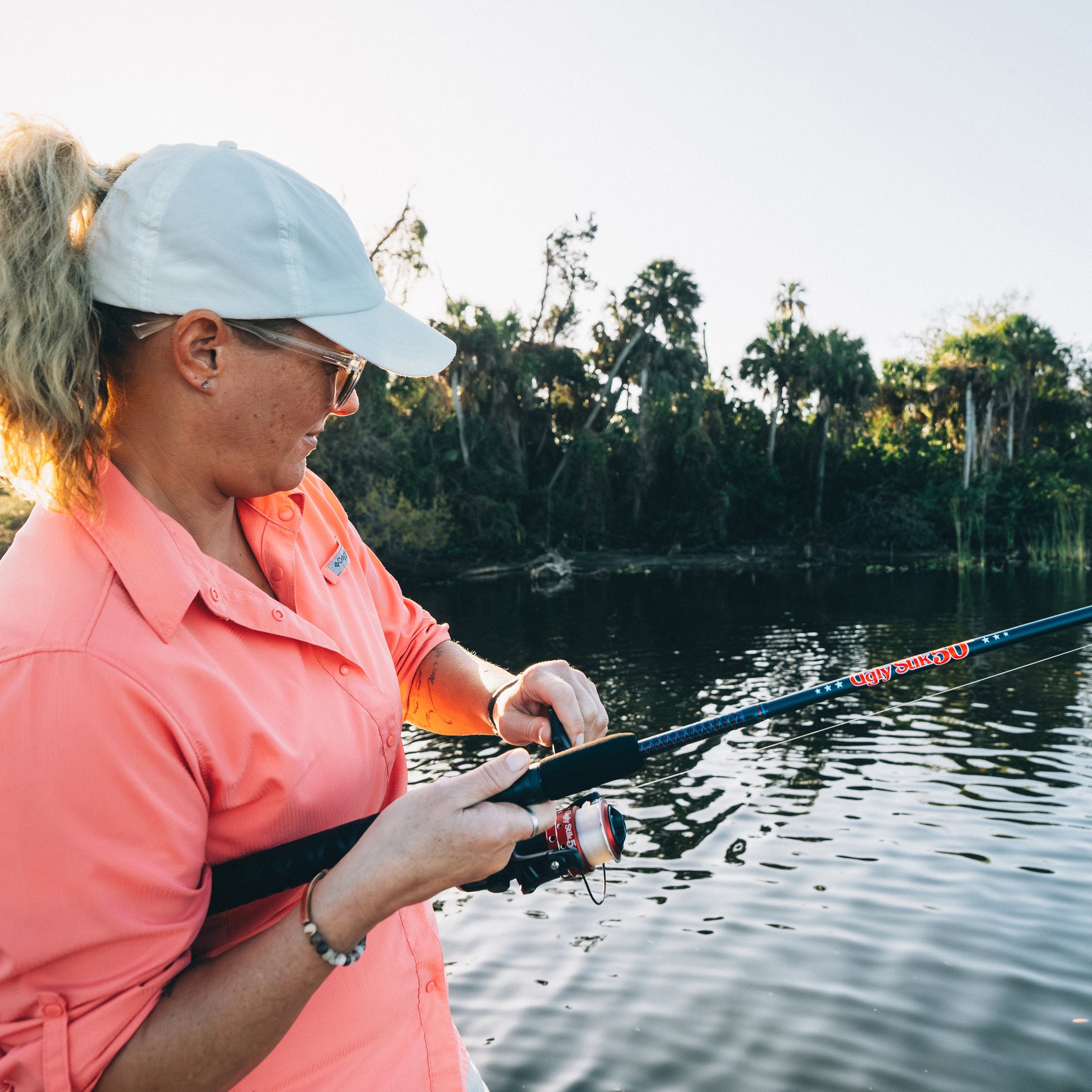Woman fishing with an Ugly Stik 50th Anniversary Spinning Combo by a lake with trees in the background