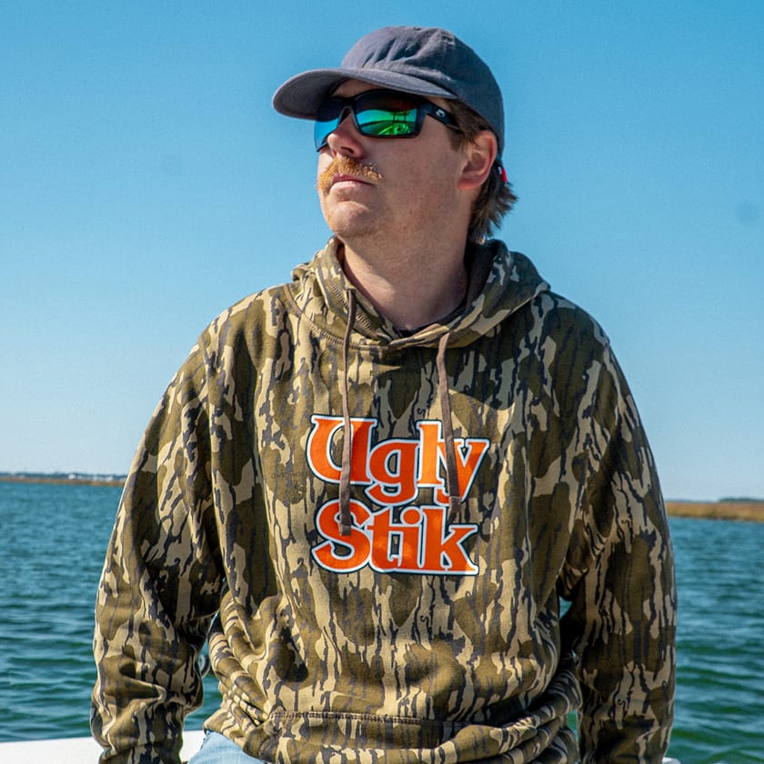 Person wearing a Mossy Oak hoodie with 'Ugly Stik' logo by a body of water.
